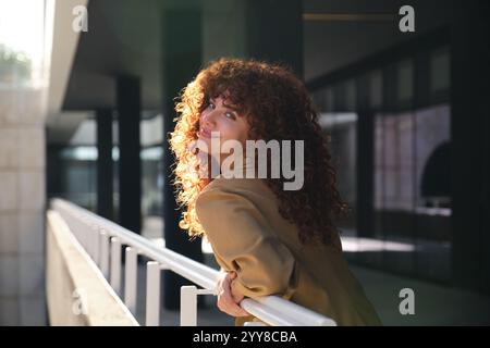 Femme d'affaires souriante avec les cheveux roux bouclés appuyés sur la rampe Banque D'Images
