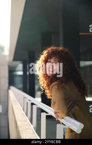 Femme d'affaires rousse souriante s'appuyant sur la rampe à l'extérieur du bureau Banque D'Images