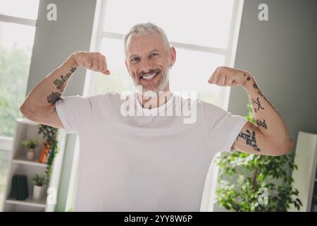 Homme âgé avec des tatouages fléchissant les muscles en t-shirt blanc dans un cadre intérieur lumineux Banque D'Images