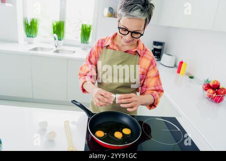 Charmante femme âgée appréciant la cuisine dans une cuisine lumineuse, craquant des œufs dans une poêle pour le petit déjeuner Banque D'Images