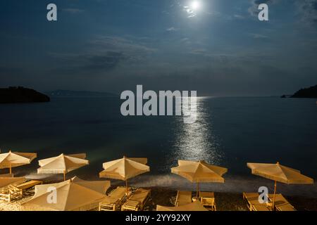 Une plage paisible au clair de lune avec des chaises longues vides et des parasols de paille sous un ciel nocturne éclatant. Le clair de lune se reflète sur la mer calme, créant un Ser Banque D'Images