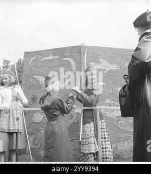 Les enfants dans les années 1940 Deux filles attrapent quelque chose dans le jeu classique de l'étang à poissons. Une ficelle avec une pince à linge au lieu d'un hameçon est attachée à un bâton qui est censé représenter une canne à pêche. La jeune fille pêche alors dans l'étang à poissons avec la pince à linge de l'autre côté de l'écran qui n'est pas visible. Derrière l'écran, quelqu'un attache quelque chose à la pince à linge, généralement un sac de bonbons. Quand la fille sent un remorqueur comme si un poisson mord, elle tire la canne à pêche et obtient les bonbons. Un plaisir simple et inoffensif qui était commun dans le passé. Suède 1949. Kristoffersson réf. A Banque D'Images