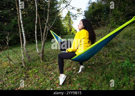 Femme aux cheveux bouclés portant une veste jaune travaillant sur son ordinateur portable tout en étant assise sur un hamac parmi les arbres dans la forêt, mettant en vedette le travail à distance et le digi Banque D'Images