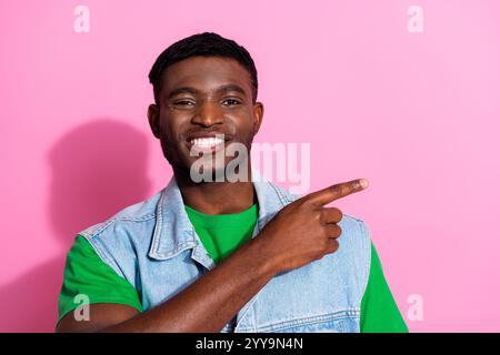 Un jeune homme élégant dans un gilet en denim tendance pointe avec confiance sur le côté, debout sur un fond rose vif Banque D'Images