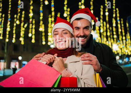 Couple souriant portant des chapeaux de Père Noël tenant des sacs à provisions pendant la nuit festive de Noël, profitant de l'esprit de vacances avec des lumières, montrant le bonheur et l'excitation tout en célébrant la saison des dons. Photo de haute qualité Banque D'Images