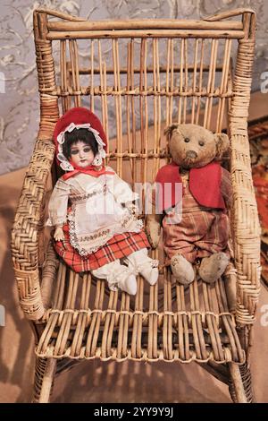 Poupée en porcelaine vintage vêtue de vêtements traditionnels de la fin du 19ème siècle assis à côté d'un ours en peluche usé portant un gilet rouge sur cha en osier rustique Banque D'Images