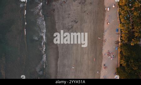 Vue aérienne d'une plage tropicale au coucher du soleil avec des palmiers, rivage rocheux, et les visiteurs dispersés appréciant le paysage côtier serein. Banque D'Images