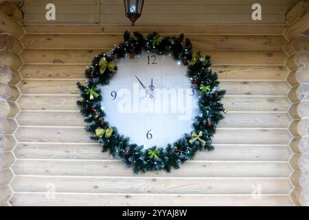 Une grande horloge est montée sur un mur en bois, entourée d'une couronne de Noël festive. La couronne présente du feuillage vert, des rubans et des ornements, de la créatine Banque D'Images