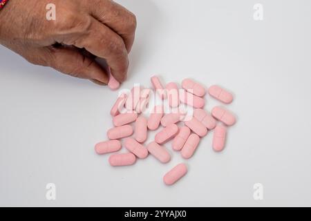 Plan de dessus d'une main senior ramassant des pilules roses dispersées sur une surface blanche et propre. Idéal pour une utilisation dans les domaines médical, pharmaceutique ou médical Banque D'Images