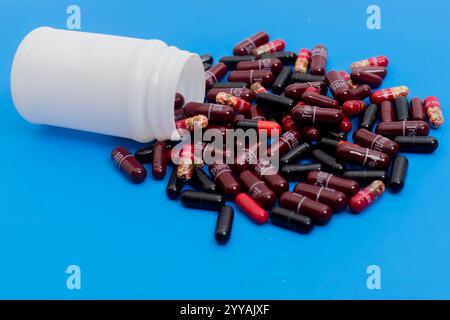 Une bouteille en plastique blanc avec des capsules de médicaments rouges et noires dispersées autour sur un fond bleu vif, soins de santé, médicaments et concept de pharmacie Banque D'Images