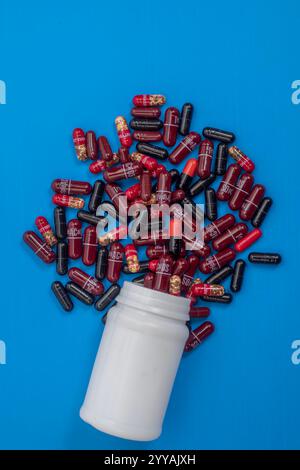 Une bouteille en plastique blanc avec des capsules de médicaments rouges et noires dispersées autour sur un fond bleu vif, soins de santé, médicaments et concept de pharmacie Banque D'Images