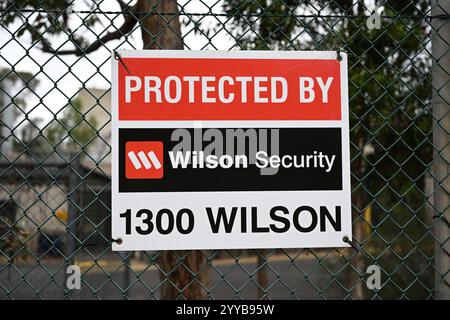 Protégé par Wilson Security signe avec le logo de l'entreprise, sur une clôture à maillons de chaîne, avec des bâtiments et des arbres en arrière-plan Banque D'Images