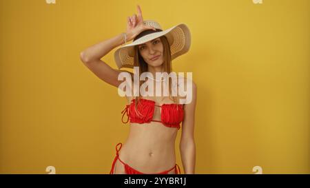 Une belle femme blonde portant un bikini rouge et un chapeau de soleil pose contre un mur isolé de fond jaune avec une expression ludique, mettant en valeur son R Banque D'Images