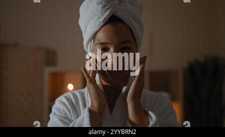 Femme appliquant de la crème dans une salle de spa de bien-être, portant un peignoir et une serviette sur la tête, souriant et profitant d'un traitement de beauté à l'intérieur. Banque D'Images