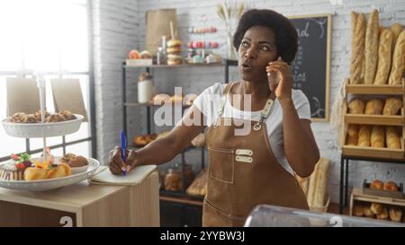 Femme parlant au téléphone et prenant des notes dans une boulangerie avec du pain et des pâtisseries affichées autour Banque D'Images