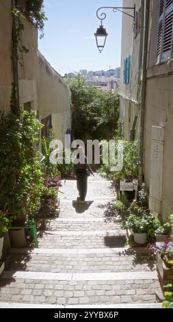 Femme explorant l'étroite ruelle européenne entourée de plantes luxuriantes sur une journée ensoleillée dans un charmant paysage urbain avec de vieilles lanternes projetant des ombres et ston historique Banque D'Images