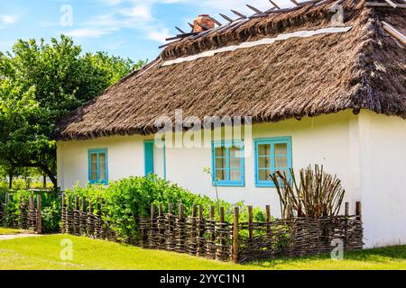 Une maison blanche avec des fenêtres bleues et un toit de chaume. La maison est entourée d'une clôture en bois et dispose d'un jardin en face Banque D'Images