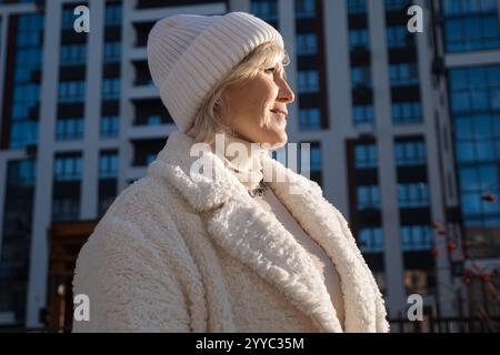 Femme âgée souriante portant des vêtements d'hiver profitant de la lumière du soleil dans un cadre urbain Banque D'Images