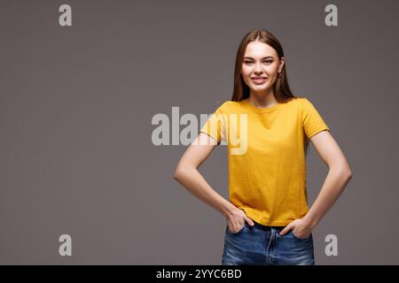 Portrait de jeune belle femme en jeans et T-shirt tenir les mains dans les poches semble isolé sur fond gris de studio Banque D'Images