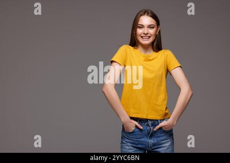 Portrait de jeune belle femme en jeans et T-shirt tenir les mains dans les poches semble isolé sur fond gris de studio Banque D'Images