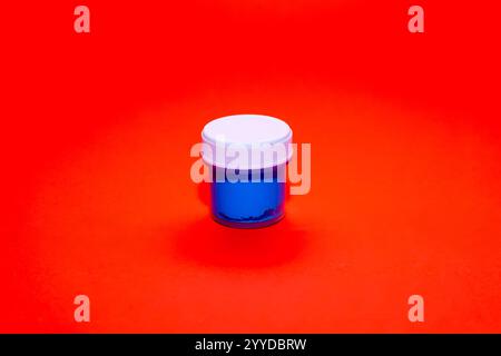 Un petit contenant de peinture bleu vif se trouve centré sur une surface rouge vif, mettant en valeur le contraste des couleurs. Le couvercle est blanc, ajoutant au visuel a Banque D'Images
