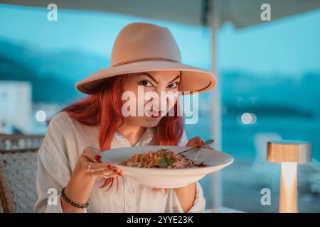 Femme rousse faisant un visage dégoûté tout en tenant une assiette de pâtes dans un restaurant au bord de la mer Banque D'Images
