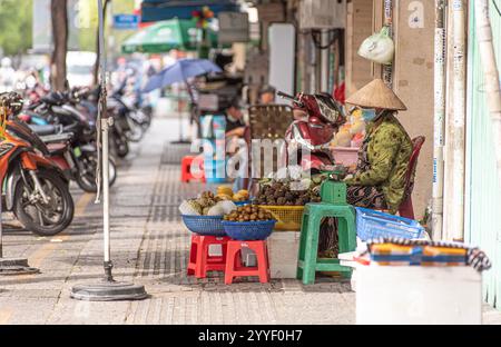 Une femme portant un masque facial est assise sur un tabouret devant un stand de fruits. Il y a plusieurs paniers de fruits sur le stand, y compris des pommes et des oranges. Banque D'Images