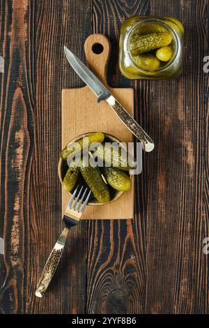 Concombres marinés dans un petit bol sur une planche à découper en bois avec un couteau et une fourchette et un pot ouvert de cornichons se trouve en arrière-plan, améliorant le Banque D'Images