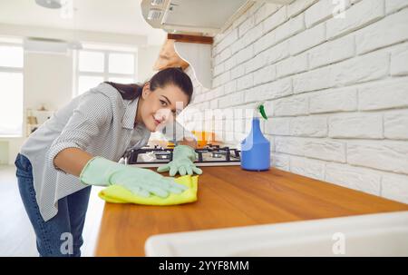 Heureuse femme au foyer souriante portant des gants de protection en caoutchouc dans une table de travail confortable de nettoyage de cuisine Banque D'Images