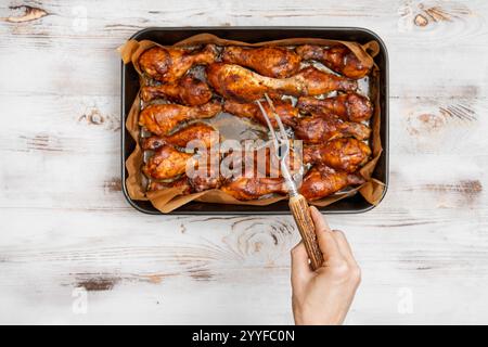 Une main tient une fourchette pour servir des pilons de poulet croustillants cuits au four disposés sur un plateau. La configuration est sur une table en bois rustique, créant une atmosphère de salle à manger confortable Banque D'Images