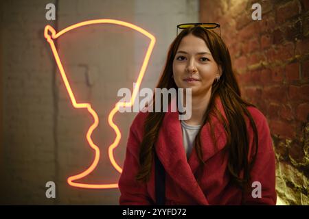Une jeune femme aux longs cheveux bruns sourit tout en portant un manteau rose, assise dans un endroit tendance orné de néon vibrants L'atmosphère chaleureuse de Banque D'Images