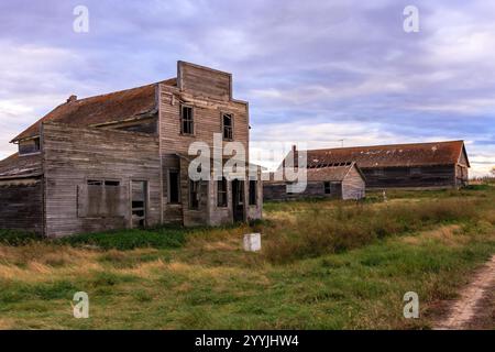 Une petite maison délabrée se trouve dans un champ avec quelques autres maisons en arrière-plan. Le ciel est nuageux et l'atmosphère est calme et paisible Banque D'Images