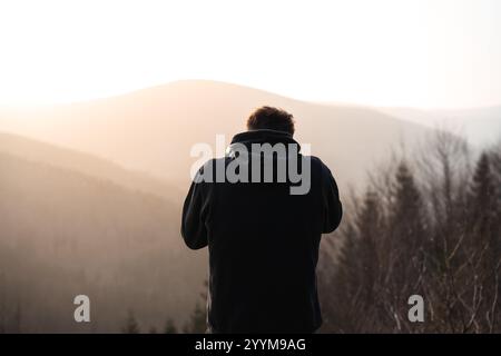 Un homme portant un sweat à capuche noir se tient en toute confiance devant une chaîne de montagnes majestueuse qui est magnifiquement capturée dans la lumière du matin Banque D'Images
