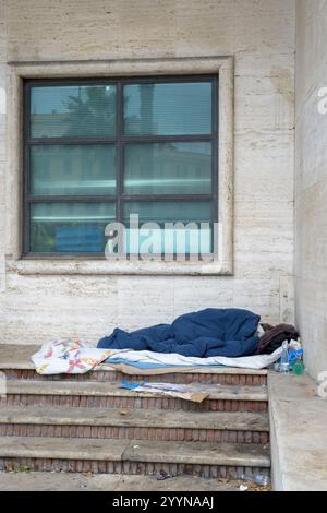 Rome, Italie. 22 décembre 2024. Un sans-abri dort dans la chaleur de son lit sous un auvent à l'abri du vent et de la pluie, tandis que le bâtiment en face se reflète dans le verre au-dessus, tard un dimanche matin de décembre à Rome. Selon le rapport du marché immobilier sur les ventes notariées et les prêts hypothécaires, publié le 19 décembre 2024 par l'Institut National de la Statistique (ISTAT), ''au premier trimestre 2024, après une tendance sensiblement stable observée au quatrième trimestre 2023, les transactions immobilières ont repris la tendance négative observée à partir du troisième trimestre 2022. T Banque D'Images