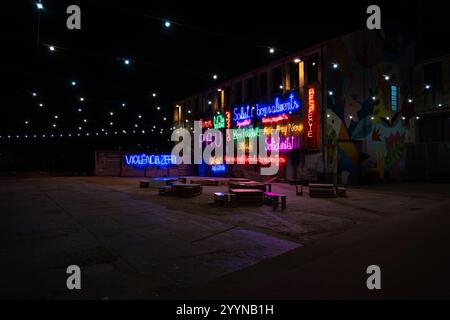 Place de ville avec chaises et tables en bois recyclé illuminées par des lettres néon colorées promouvant la paix et la solidarité et des lumières suspendues Banque D'Images