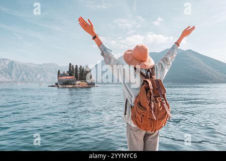 Touriste femme portant sac à dos et chapeau levant les bras profitant de la vue panoramique de l'île avec monastère dans la baie de Kotor, Perast, Monténégro Banque D'Images