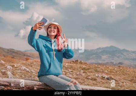 Randonneur féminin souriant prenant selfie avec smartphone tout en étant assis sur la bûche en bois dans le paysage de montagne Banque D'Images