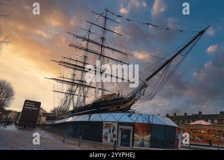 L'historique Cutty Sark est situé contre un ciel de coucher de soleil à Londres. Une structure en verre moderne et un carrousel festif soulignent la période de Noël. Banque D'Images