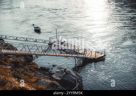 Une scène paisible au bord de la rivière avec un quai en bois s'étendant dans les eaux calmes, un petit bateau amarré à proximité, et des reflets ensoleillés créant un calme à Banque D'Images