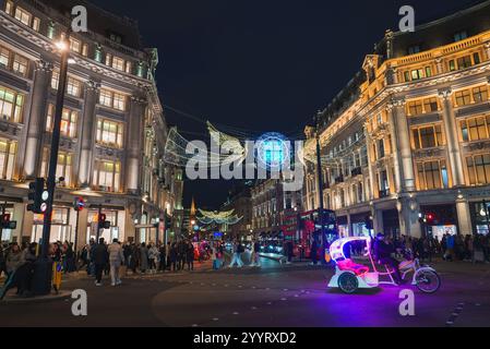 Oxford Circus à Londres présente des lumières festives, y compris un globe et des ailes d'ange. Des bus à impériale rouges et un pédicab rehaussent la scène animée. Banque D'Images