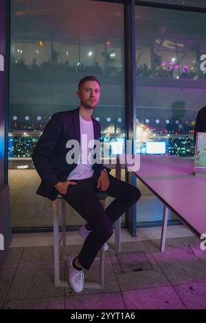 Un homme est assis sur un tabouret dans un intérieur moderne de grande hauteur avec de grandes fenêtres, mettant en valeur le paysage urbain illuminé de Londres la nuit pendant la saison de Noël Banque D'Images