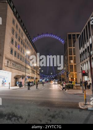 Le London Eye brille en violet sur fond de bâtiments modernes et classiques. Les piétons et les véhicules ajoutent de la vie à la scène nocturne festive. Banque D'Images