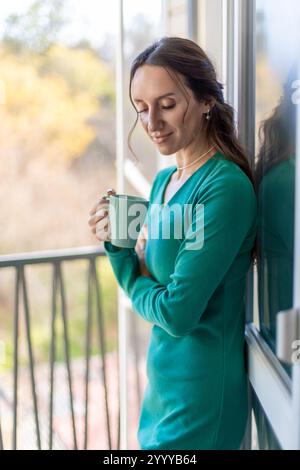 Femme en robe à manches longues sarcelle se tient sur le balcon, tenant une tasse sarcelle, regardant vers le bas. La lumière du soleil l'éclaire par derrière. Banque D'Images