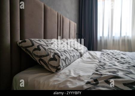 Cette chambre comprend un lit doté d'une tête de lit rembourrée, de deux oreillers à motifs de coeur et d'un couvre-lit blanc et gris. La lumière naturelle traverse le cube transparent Banque D'Images