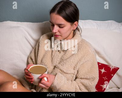 Atmosphère sereine et réconfortante est créée comme une jeune femme profite d'une matinée paisible au lit, enveloppée dans une veste sherpa beige confortable et savourant un Banque D'Images