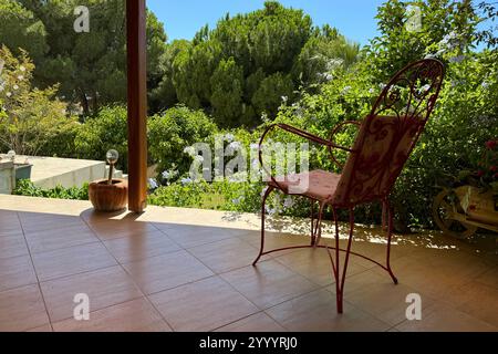 Chaise rouge classique en fer forgé et oreillers roses à motifs roses rouges de style ancien sur la terrasse de la villa de vacances dans le jardin, arbres, fleurs, ciel bleu Banque D'Images