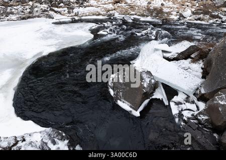 Une rivière coule à travers un terrain rocheux, partiellement recouvert de glace et de neige, mettant en valeur le froid de l'hiver. L'eau claire contraste avec l'environnement givré Banque D'Images