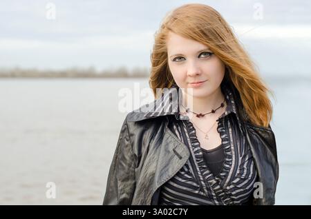 Jeune femme aux cheveux roux et à la peau claire se tient près du lac calme. Portant une veste en cuir noir et une chemise rayée, elle regarde doucement dans la caméra. Couvert s Banque D'Images