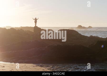 Quelqu'un en silhouette se tient au sommet de grands rochers avec les bras en l'air au coucher du soleil regardant les vagues et la mer à Essaouria, Maroc. Février/mars 2025 Banque D'Images