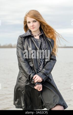 Jeune femme aux cheveux roux et à la peau claire se tient près du lac calme. Portant une veste en cuir noir et une chemise rayée, elle regarde doucement dans la caméra. Couvert s Banque D'Images